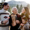A family of four outdoors during golden hour with a scenic mountainous backdrop. The elderly man holds a box of 'Bison Jerky Sticks' and individual jerky sticks. To his left, a young man with a geometric sweater holds a box of jerky. To his right, a young woman with long dark hair smiles at the elderly man, and a young boy eats a jerky stick