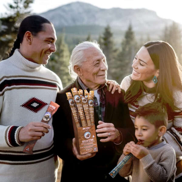 A family of four outdoors during golden hour with a scenic mountainous backdrop. The elderly man holds a box of 'Bison Jerky Sticks' and individual jerky sticks. To his left, a young man with a geometric sweater holds a box of jerky. To his right, a young woman with long dark hair smiles at the elderly man, and a young boy eats a jerky stick